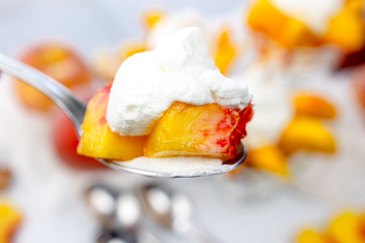 Close up view of a spoon lifting a bite of peach with whipped cream on top.