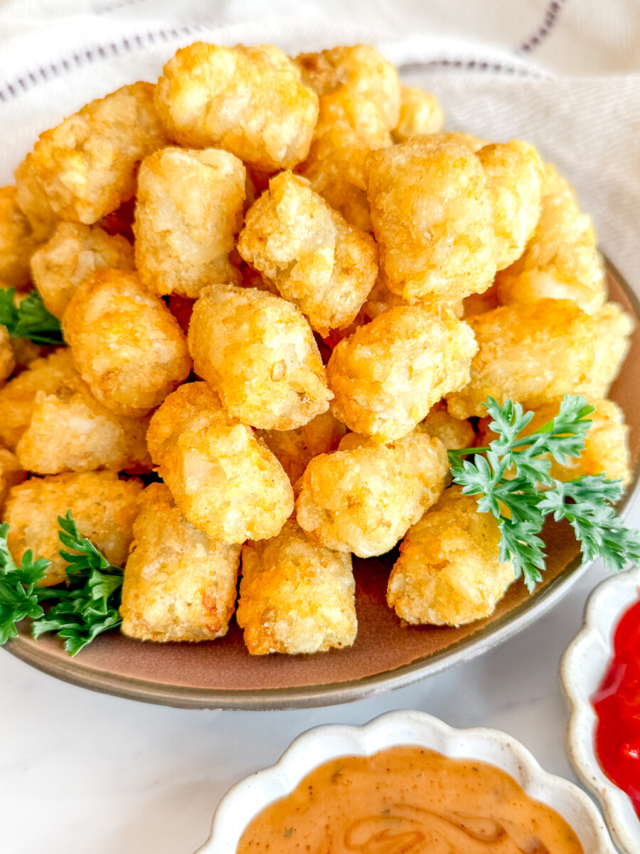 Angled view of cooked tater tots on a brown plate, garnished with parsley, next to dipping sauces in white ramekins.