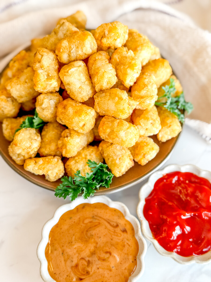 Angled view of cooked tater tots on a brown plate, garnished with parsley, next to dipping sauces in white ramekins.