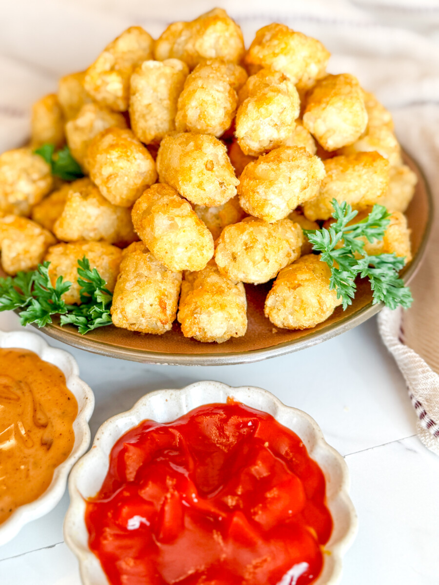 Angled view of cooked tater tots on a brown plate, garnished with parsley, next to dipping sauces in white ramekins.
