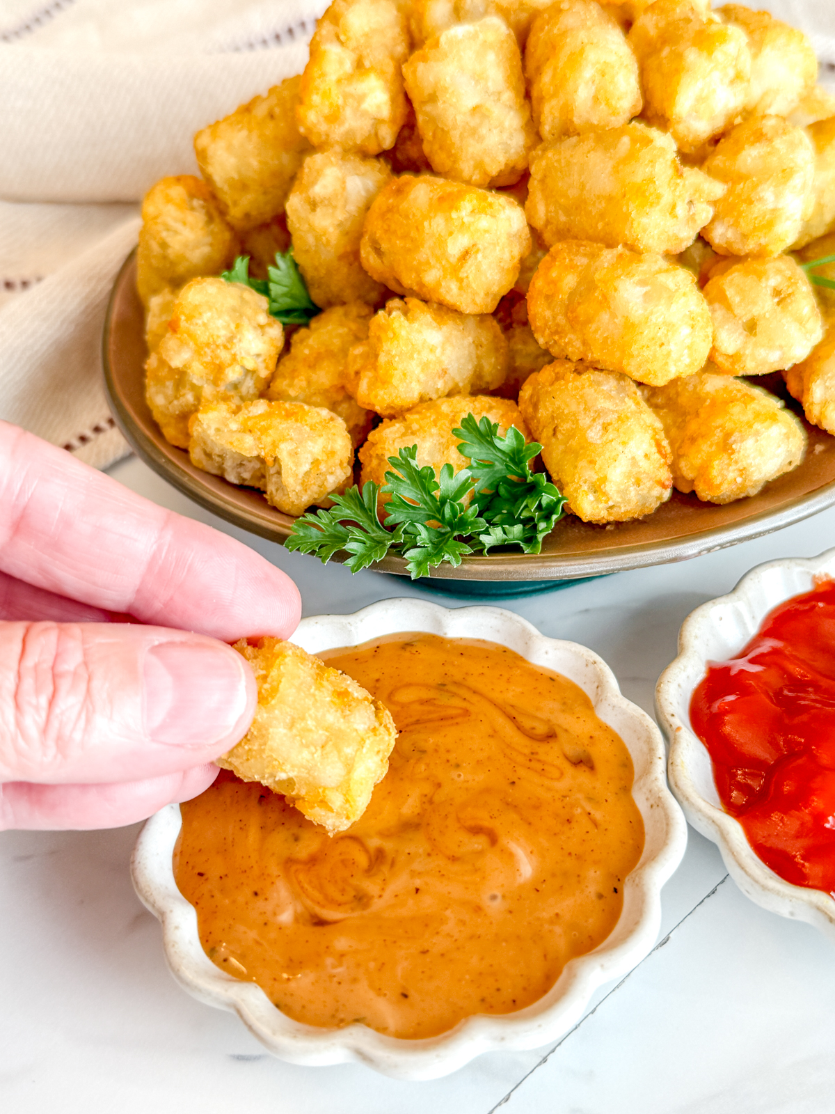 Angled view of cooked tater tots on a brown plate, garnished with parsley, next to dipping sauces in white ramekins. A hand is dipping a tater tot into some sauce.