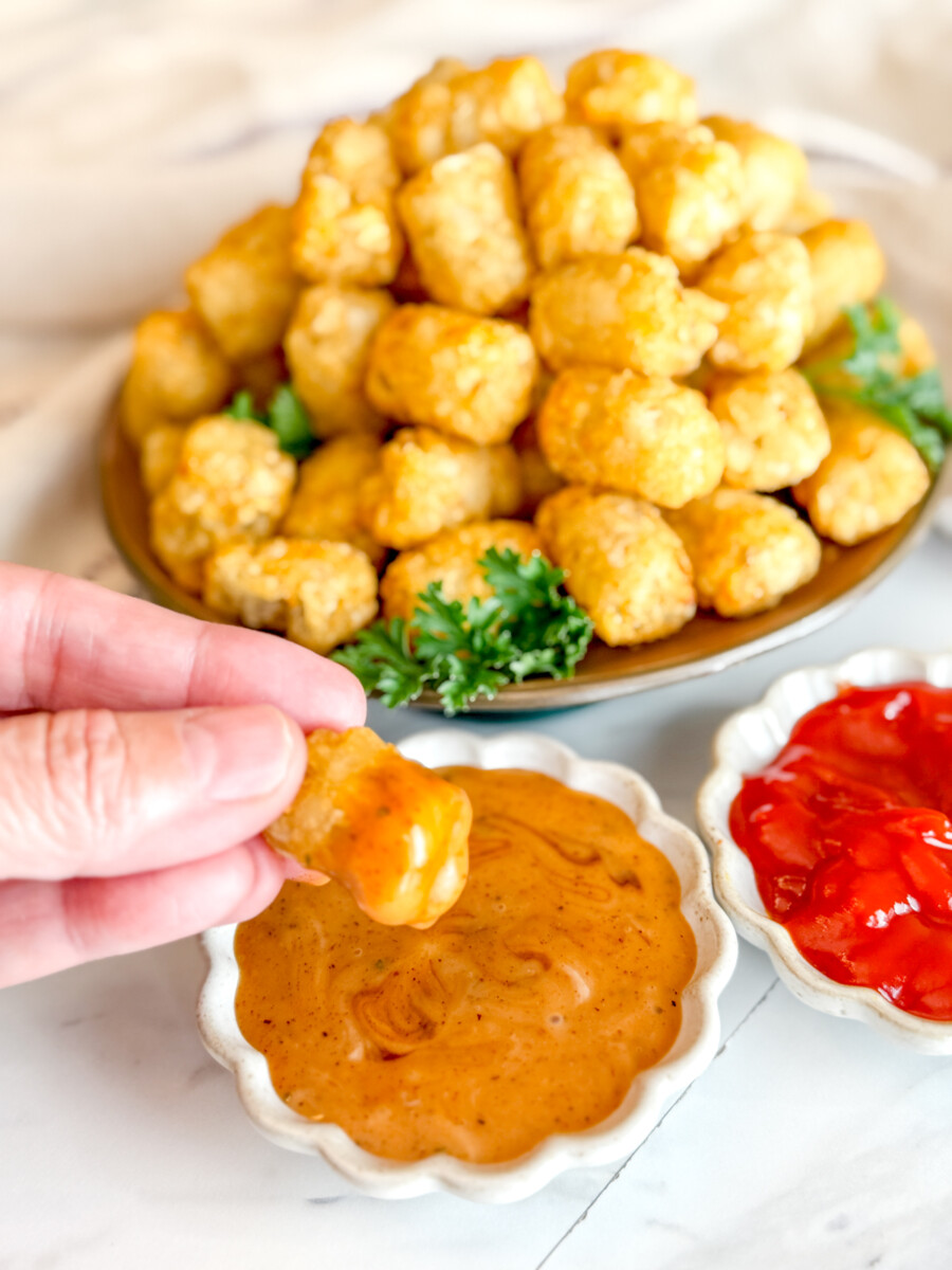 Angled view of cooked tater tots on a brown plate, garnished with parsley, next to dipping sauces in white ramekins. A hand is dipping a tater tot into some sauce.