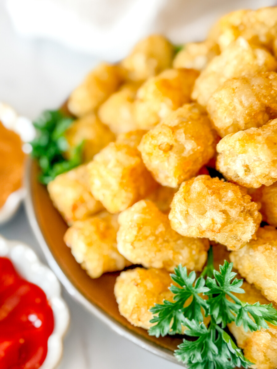 Angled view of cooked tater tots on a brown plate, garnished with parsley, next to dipping sauces in white ramekins.