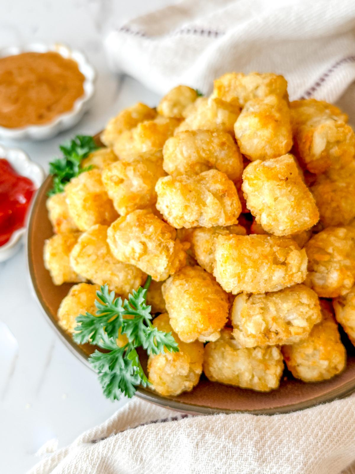 Angled view of cooked tater tots on a brown plate, garnished with parsley, next to dipping sauces in white ramekins.