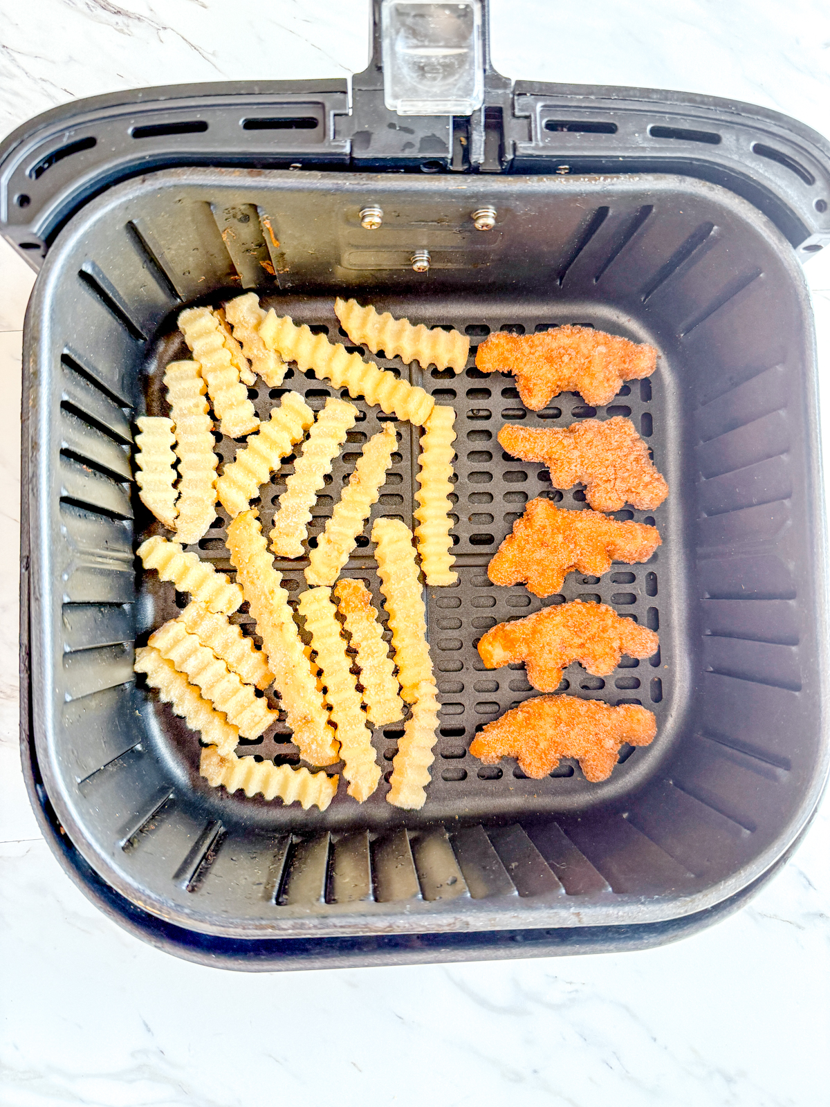 Overhead image of an air fryer basket with five dino nuggets on one half of the bottom, and a layer of crinkle cut fries on the other half.
