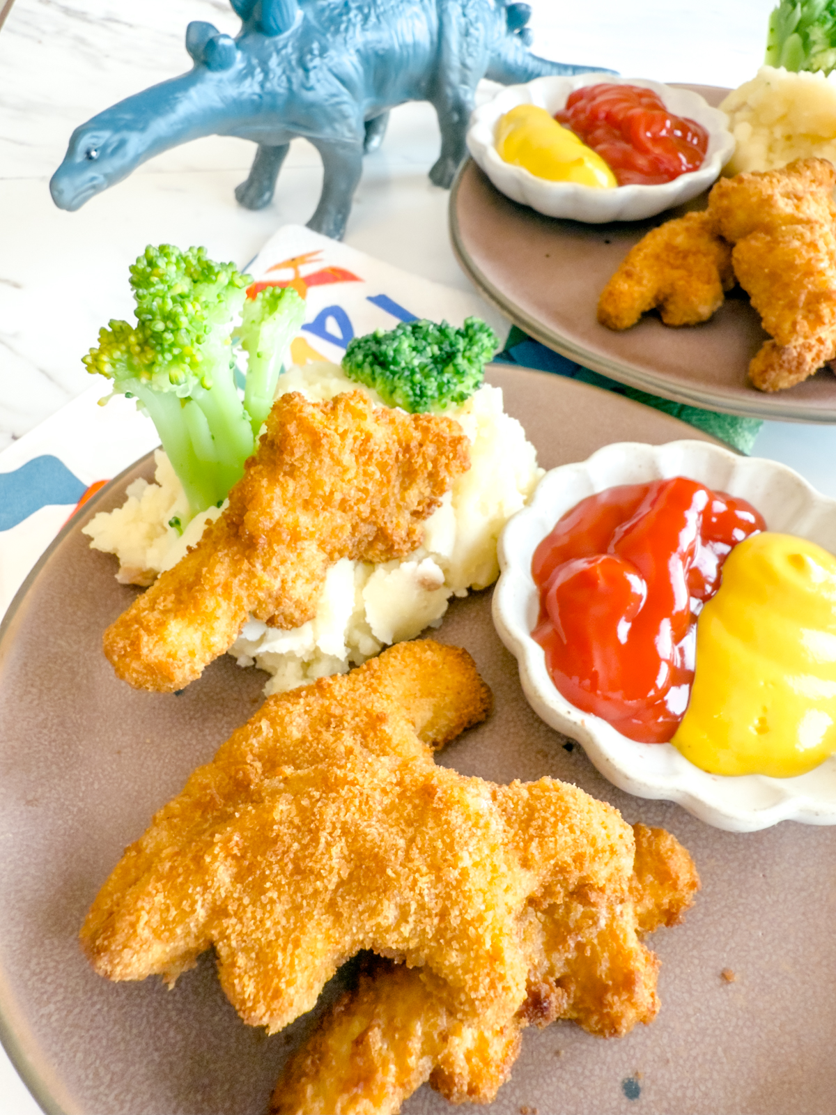 Angled view of Air Fryer Dino Nuggets on a brown plate with mashed potatoes, brocolli, and dipping sauces.