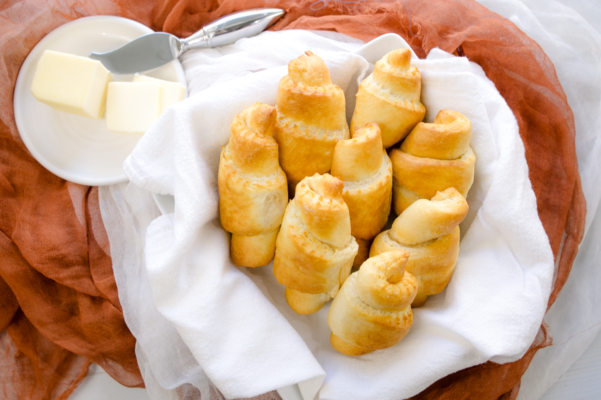 Angled view of cooked crescent rolls wrapped in a white fabric next to a small plate with butter and a knife on it.