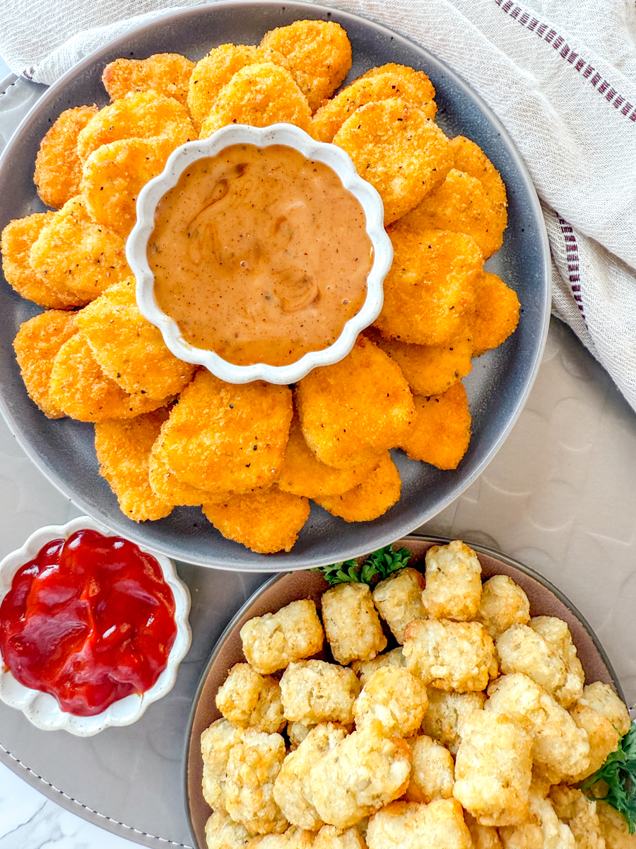 Overhead view of a plate of Air Fryer Frozen Chicken Nuggets with dipping sauces, next to a plate of tater tots.