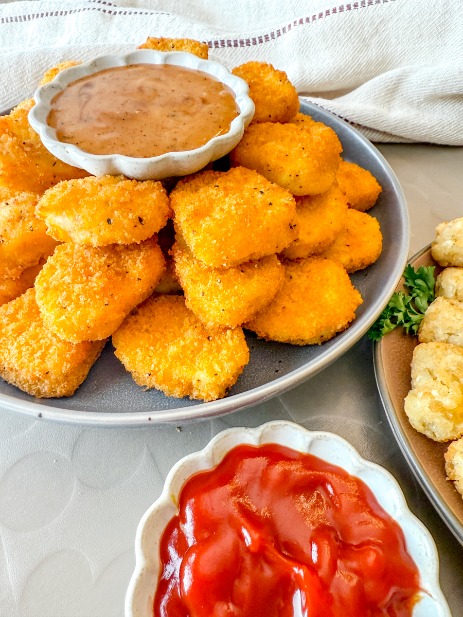 Angled view of a plate of Air Fryer Frozen Chicken Nuggets with dipping sauces, next to a plate of tater tots.