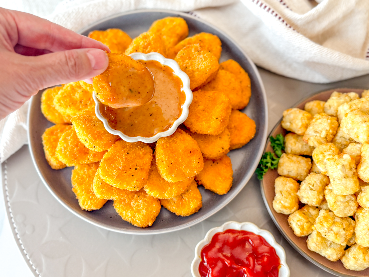 Angled view of a hand dipping a chicken nugget into a dipping sauce on a plate of Air Fryer Frozen Chicken Nuggets, next to another dipping sauce and a plate of tater tots.