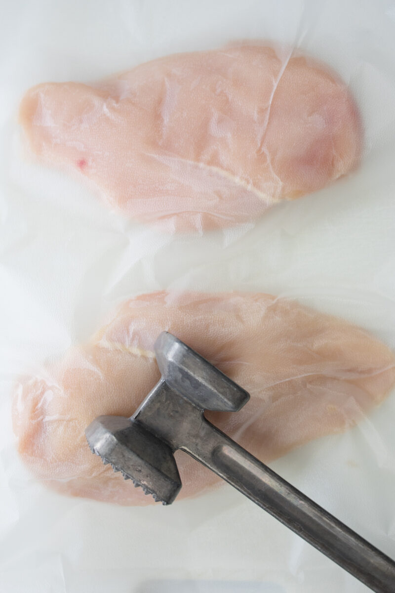 Overhead view of raw chicken breasts under plastic wrap with a tenderizing mallet on top.