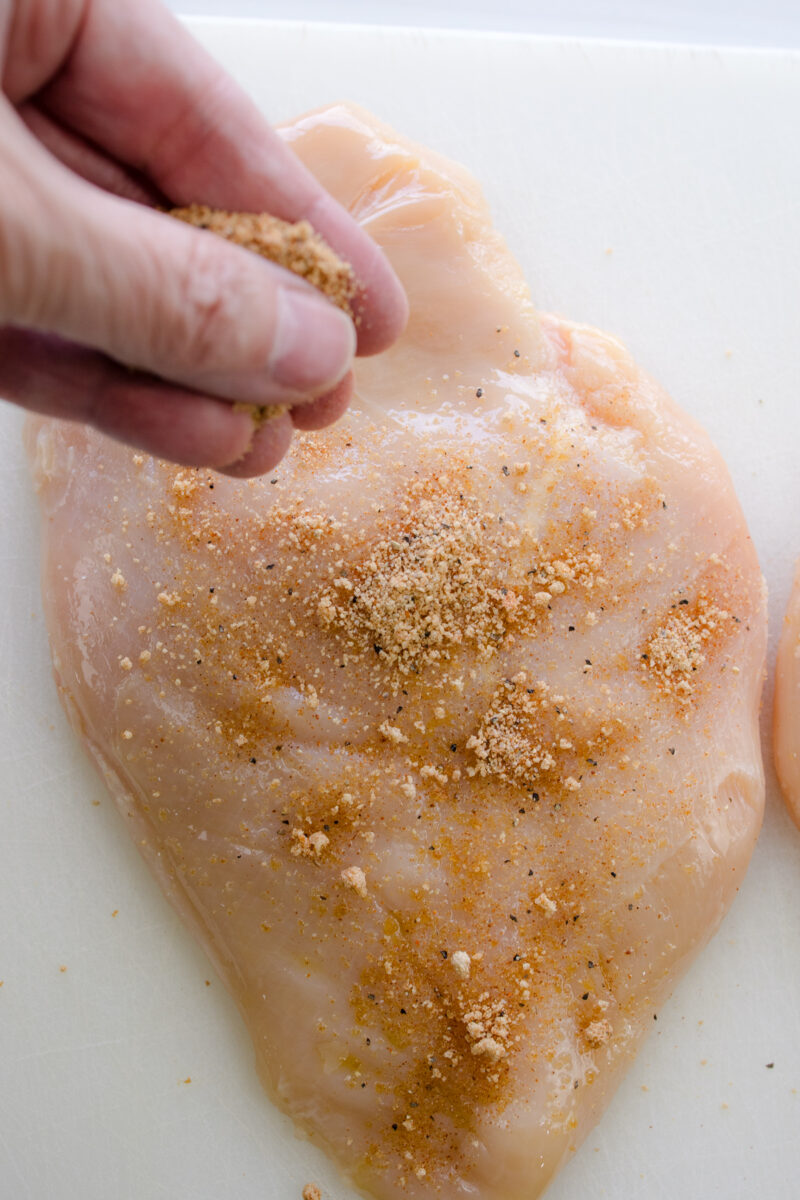 Close up overhead view of a hand sprinkling seasoning over a raw chicken breast.