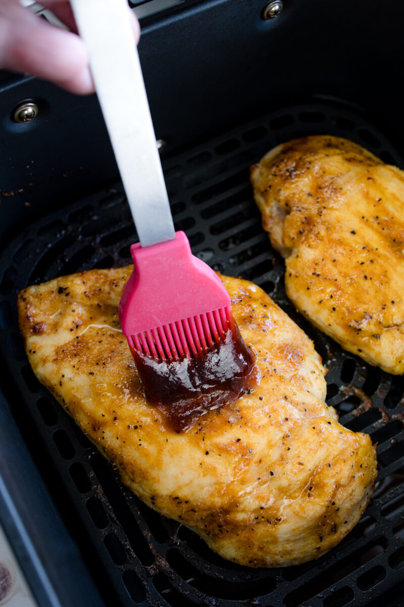 Close up view of part-cooked chicken breasts in an air fryer basket being basted with BBQ sauce on the other side after being flipped over.