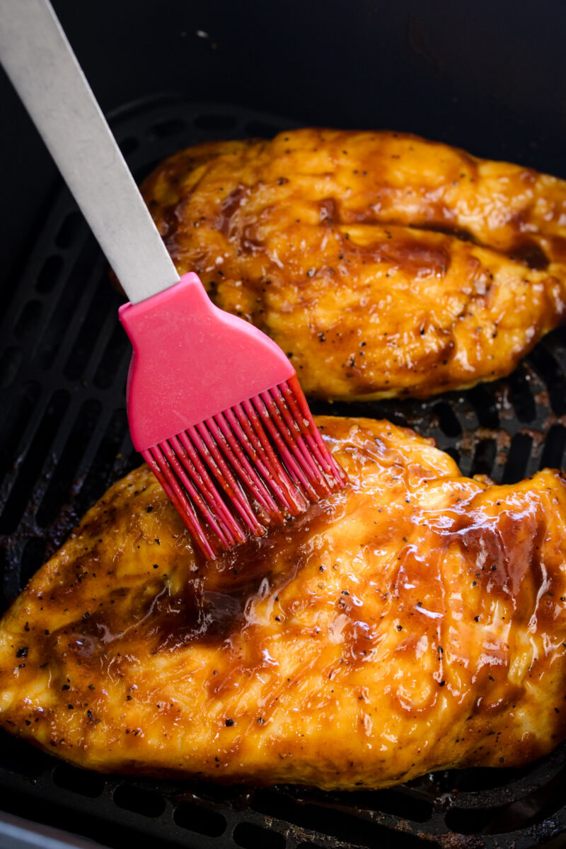 Close up view of seasoned raw chicken breasts being basted with BBQ sauce in an air fryer basket.