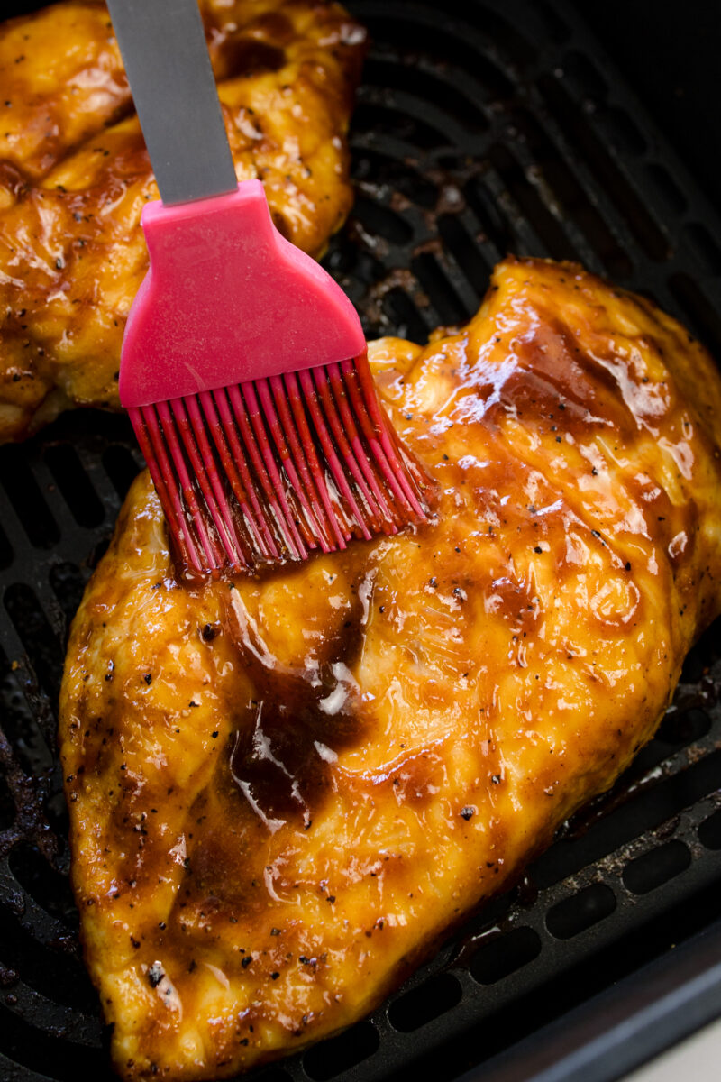 Close up view of cooked chicken breasts being basted with BBQ sauce in an air fryer basket.