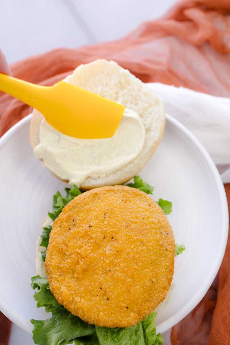 Overhead image of mayo being spread on the top half of a bun, next to an assembled chicken patty sandwich without a top, on a white plate.