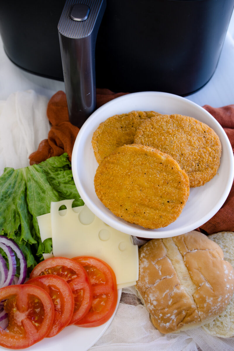 Close up image of all ingredients and toppings for making chicken patty sandwiches, next to an air fryer.