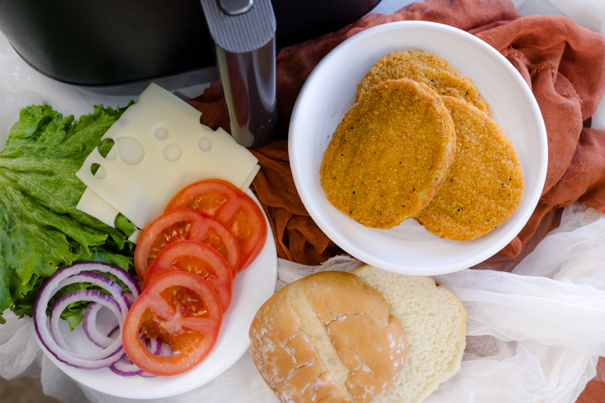 Close up image of all ingredients and toppings for making chicken patty sandwiches, next to an air fryer.