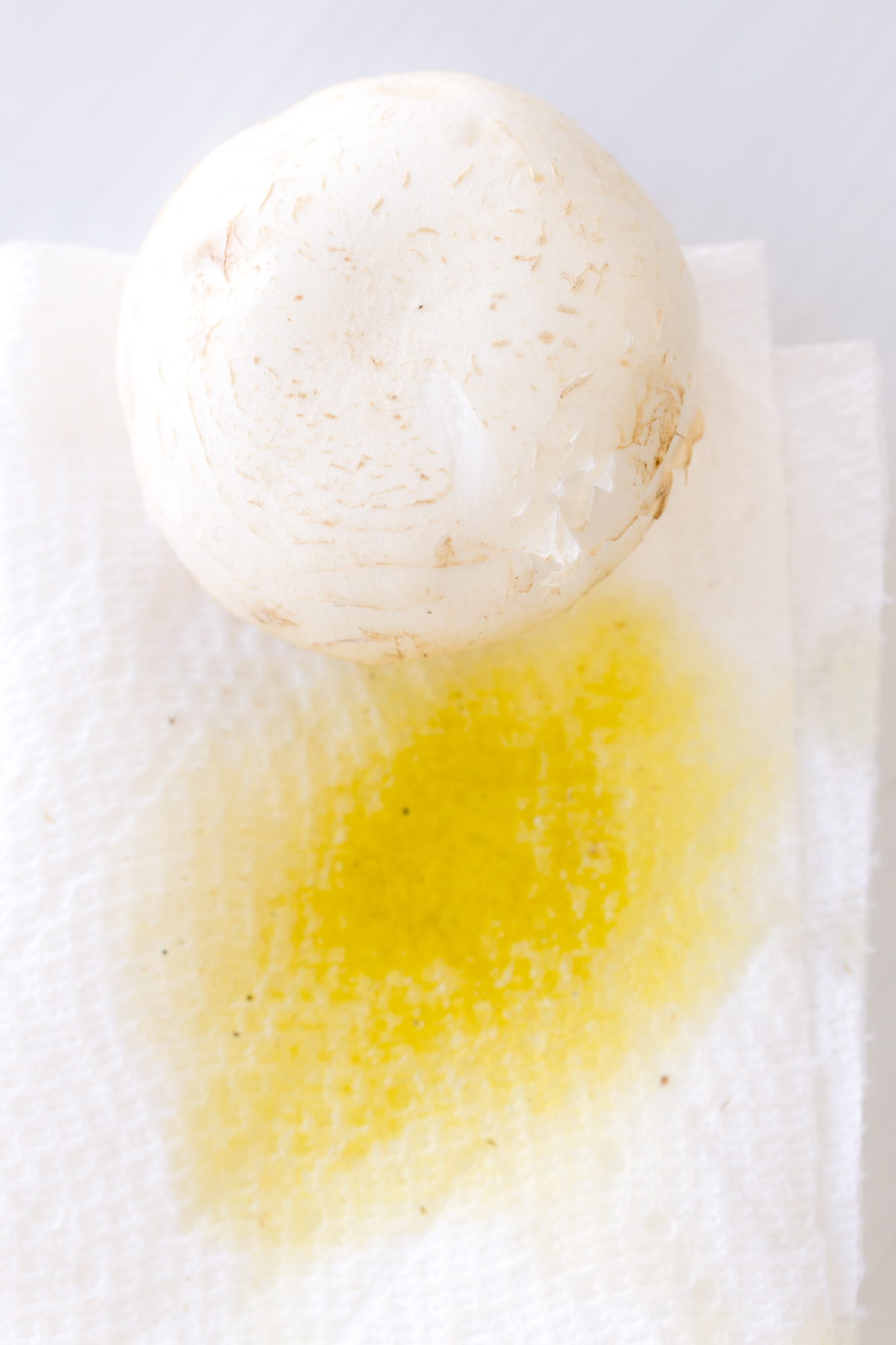 Close up image of a mushroom cap being lightly wiped with olive oil on a paper towel.