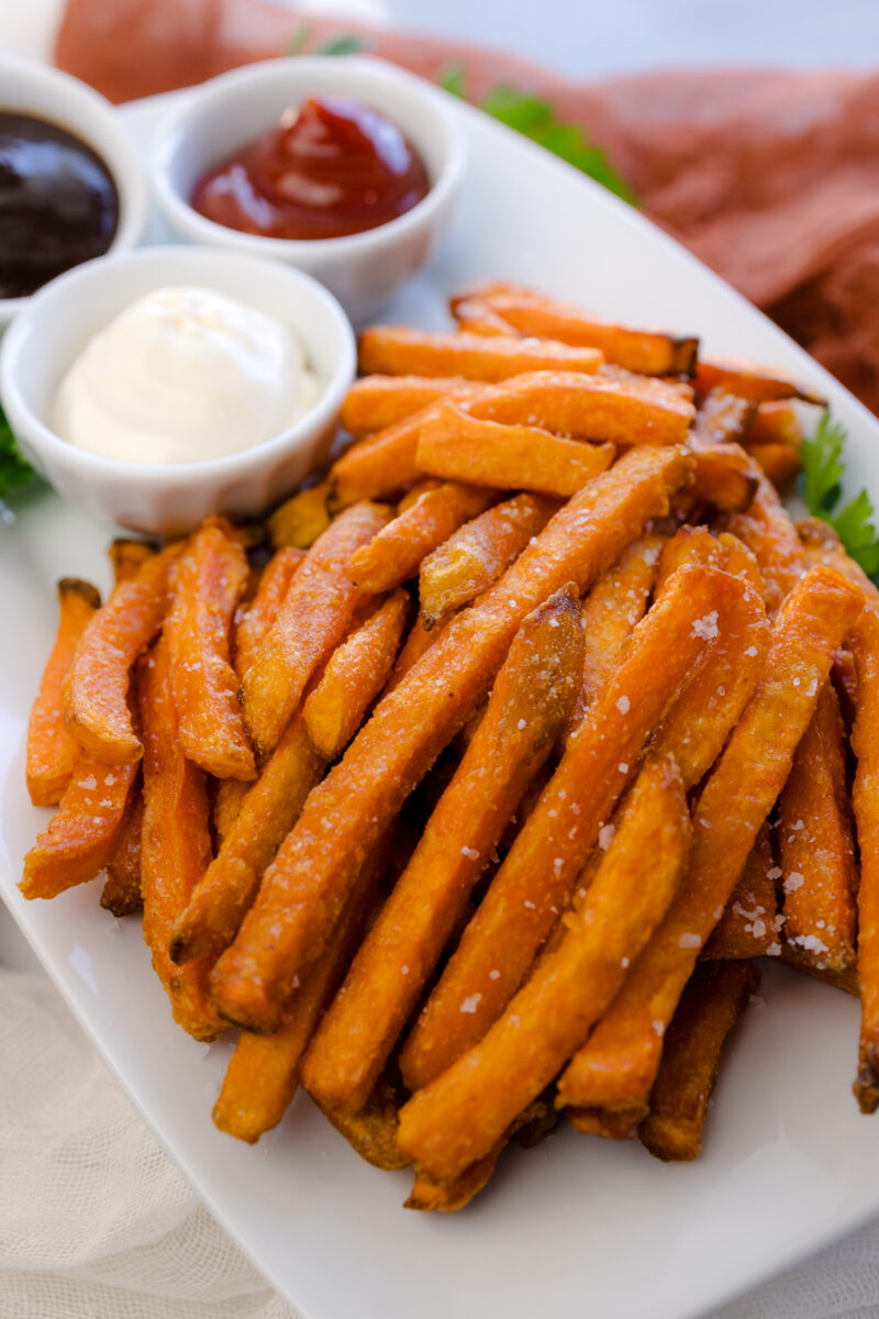 Angled view of air fried sweet potato fries on a white serving platter with dipping sauces.