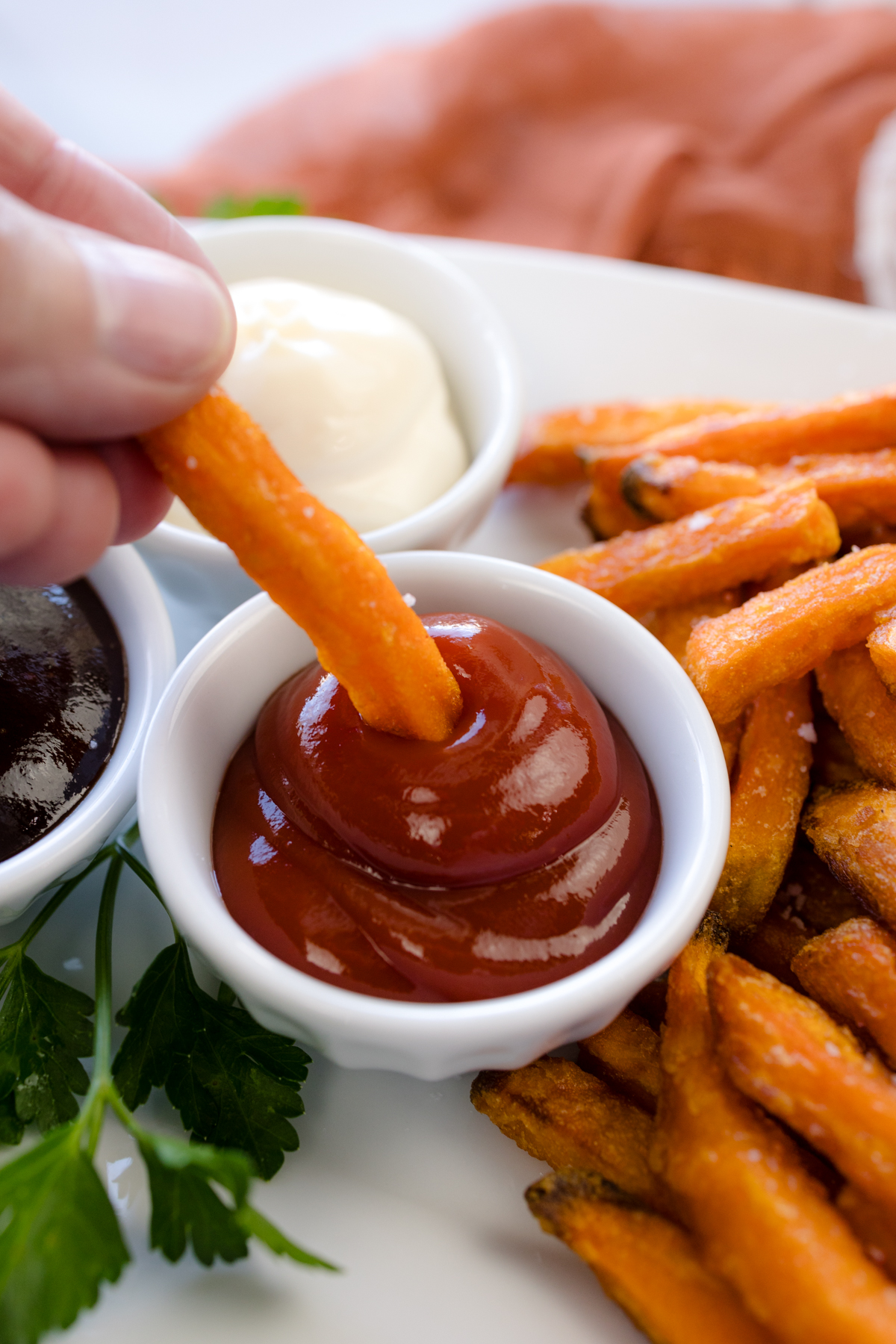Angled view of a hand dipping a sweet potato fry into ketchup, on a serving platter next to air fried sweet potato fries and other dipping sauces.