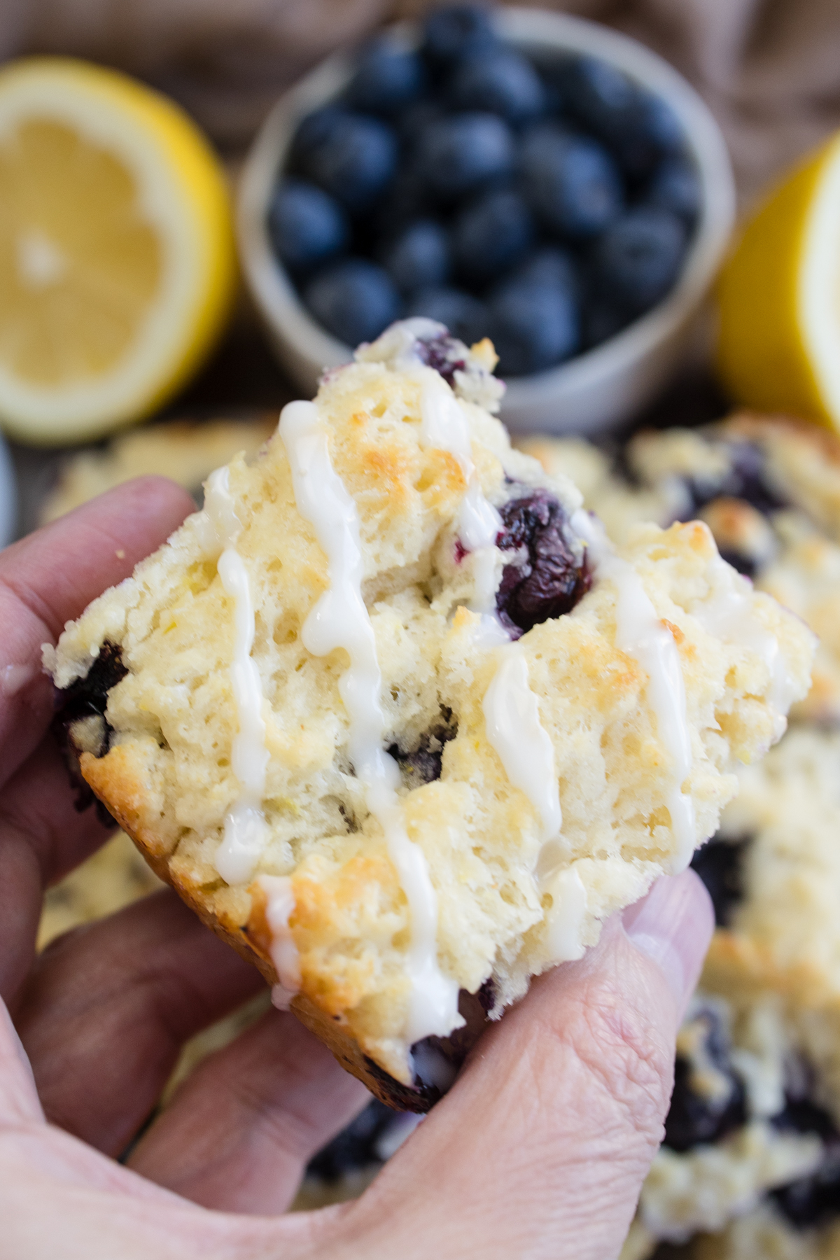 Close view of a hand holding a Blueberry Biscuit, with blueberries and half a lemon in the background.