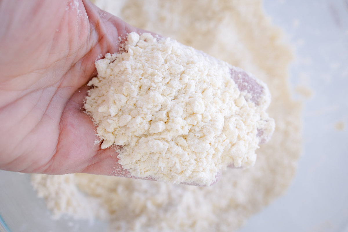 Close view of a hand holding a handful of flour over a glass bowl with flour in it.