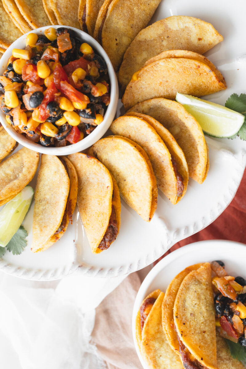 Overhead view of air fried mini tacos on a serving platter, with a few wedges of lime, all fanned out in a circle around a white bowl of salsa in the middle, next to a small plate with a few mini tacos on it with some salsa.