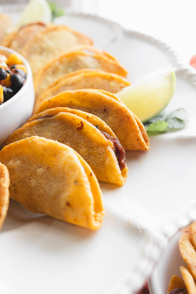 Angled view of air fried mini tacos on a serving platter, with a few wedges of lime, all fanned out in a circle around a white bowl of salsa in the middle.