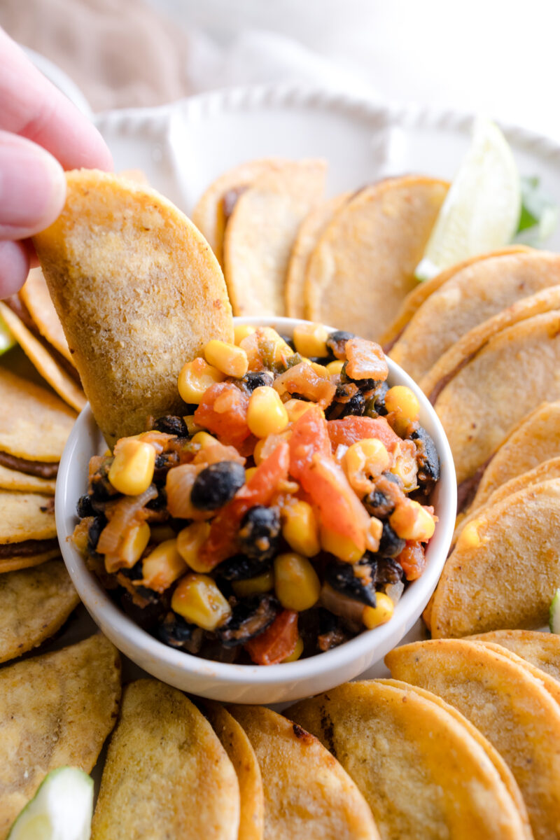 Angled view of air fried mini tacos on a serving platter, with a few wedges of lime, all fanned out in a circle around a white bowl of salsa in the middle. A hand is dipping a mini taco into the salsa bowl.