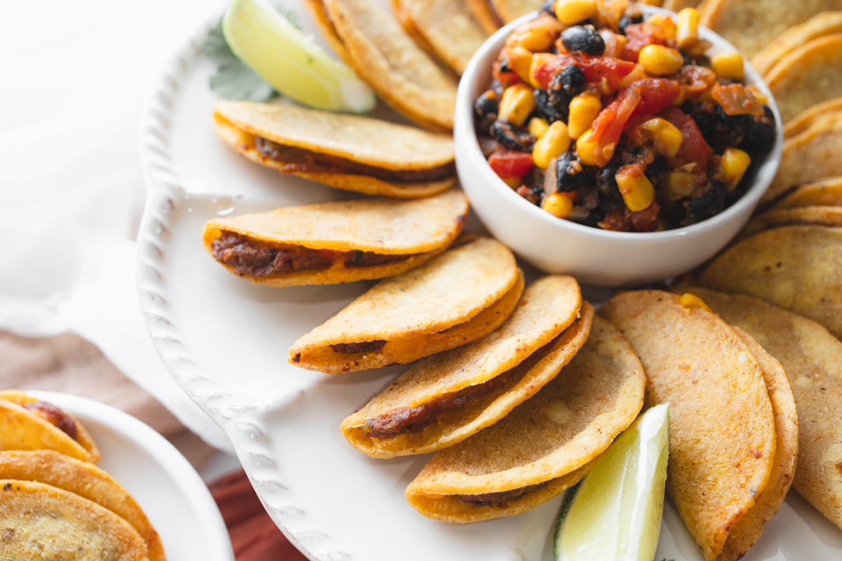 Angled view of air fried mini tacos on a serving platter, with a few wedges of lime, all fanned out in a circle around a white bowl of salsa in the middle, next to a small plate with mini tacos on it.