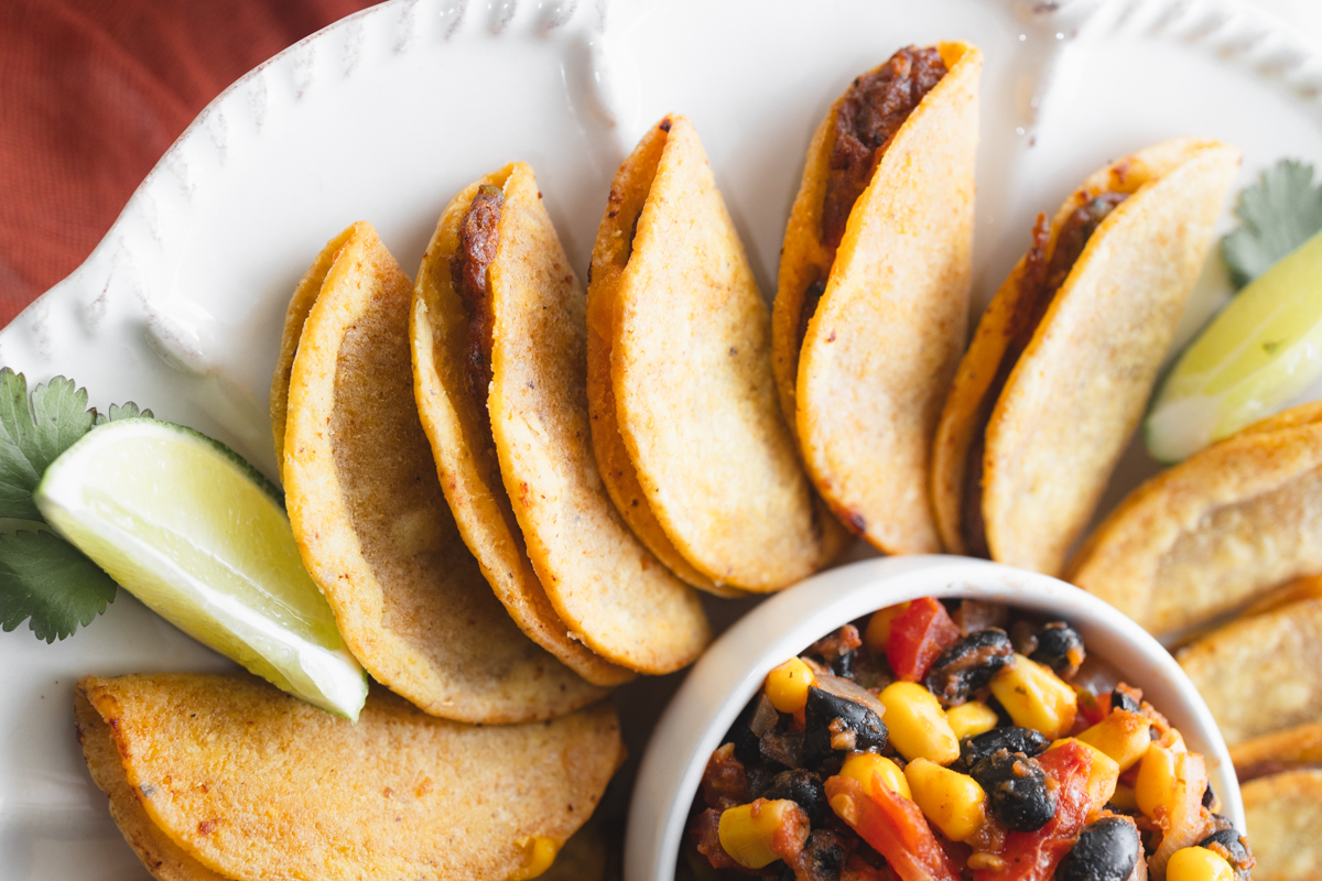 Overhead view of air fried mini tacos on a serving platter, with a few wedges of lime, all fanned out in a circle around a white bowl of salsa in the middle.