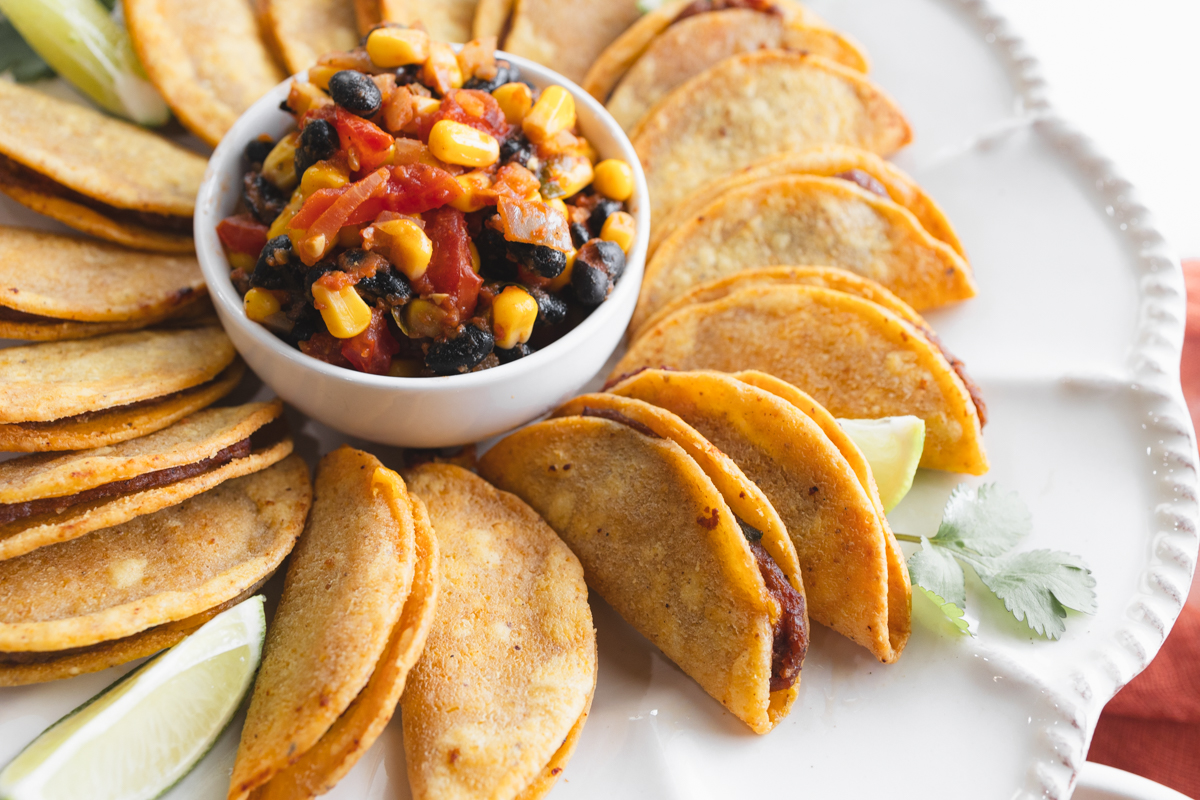 Angled view of air fried mini tacos on a serving platter, with a few wedges of lime, all fanned out in a circle around a white bowl of salsa in the middle.