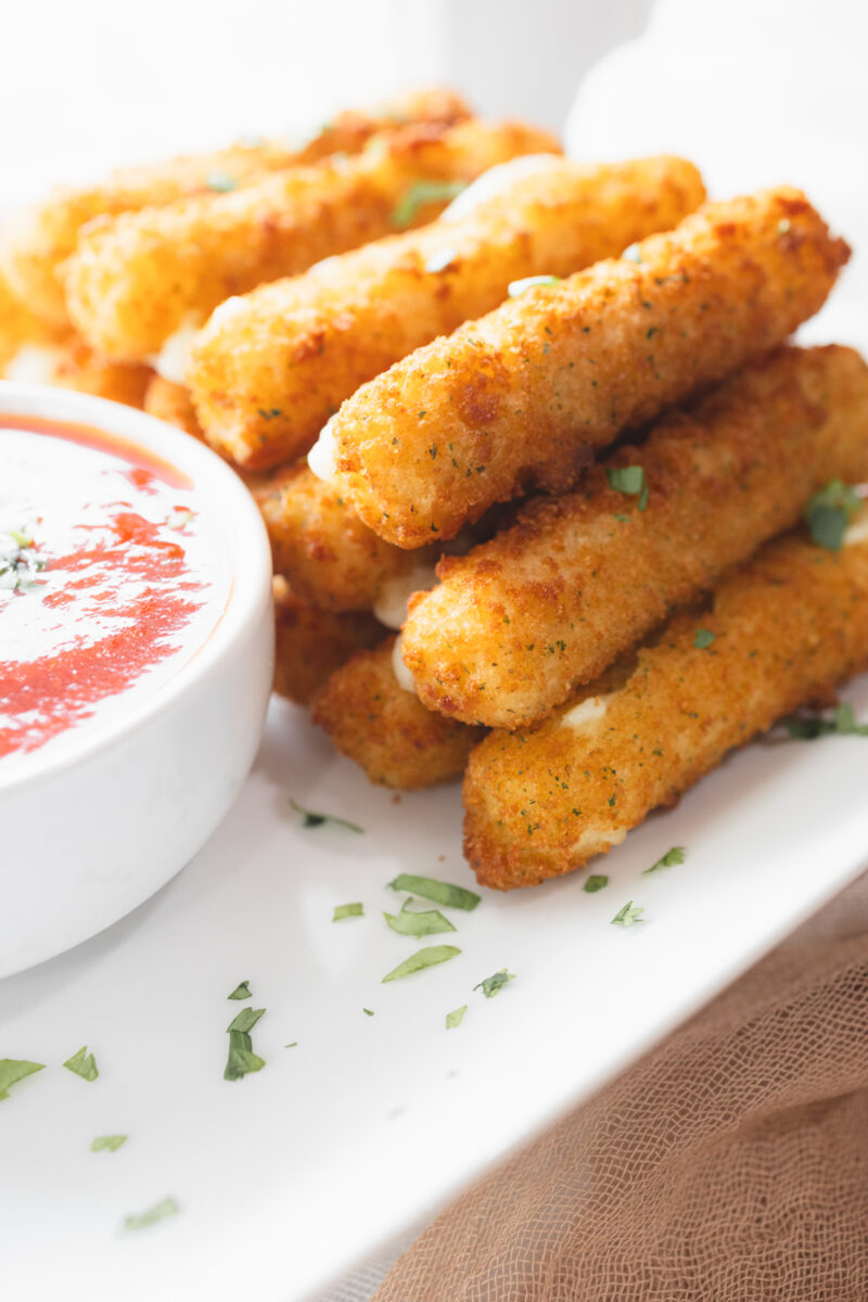 Angled view of Air Fryer Mozzarella Sticks next to a red dipping sauce in a white dipping bowl with a mozzarella stick dipped in it.