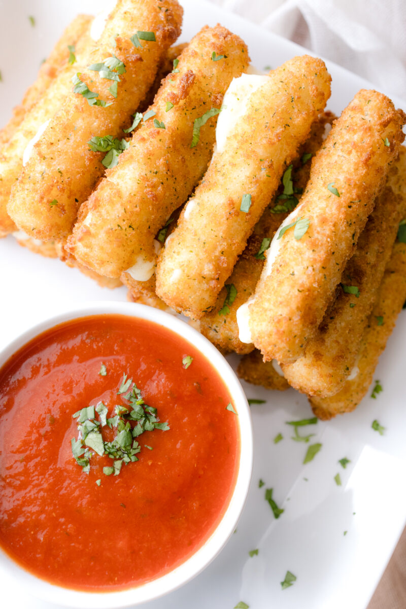 Close up overhead image of Air Fryer Mozzarella Sticks next to a red dipping sauce in a white dipping bowl.