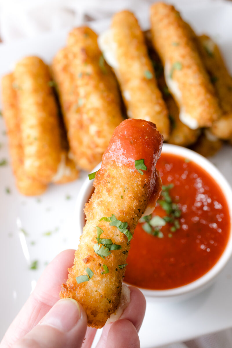 Close up angled image of Air Fryer Mozzarella Sticks next to a red dipping sauce in a white dipping bowl, with a hand holding a dipped mozzarella stick in the foreground.