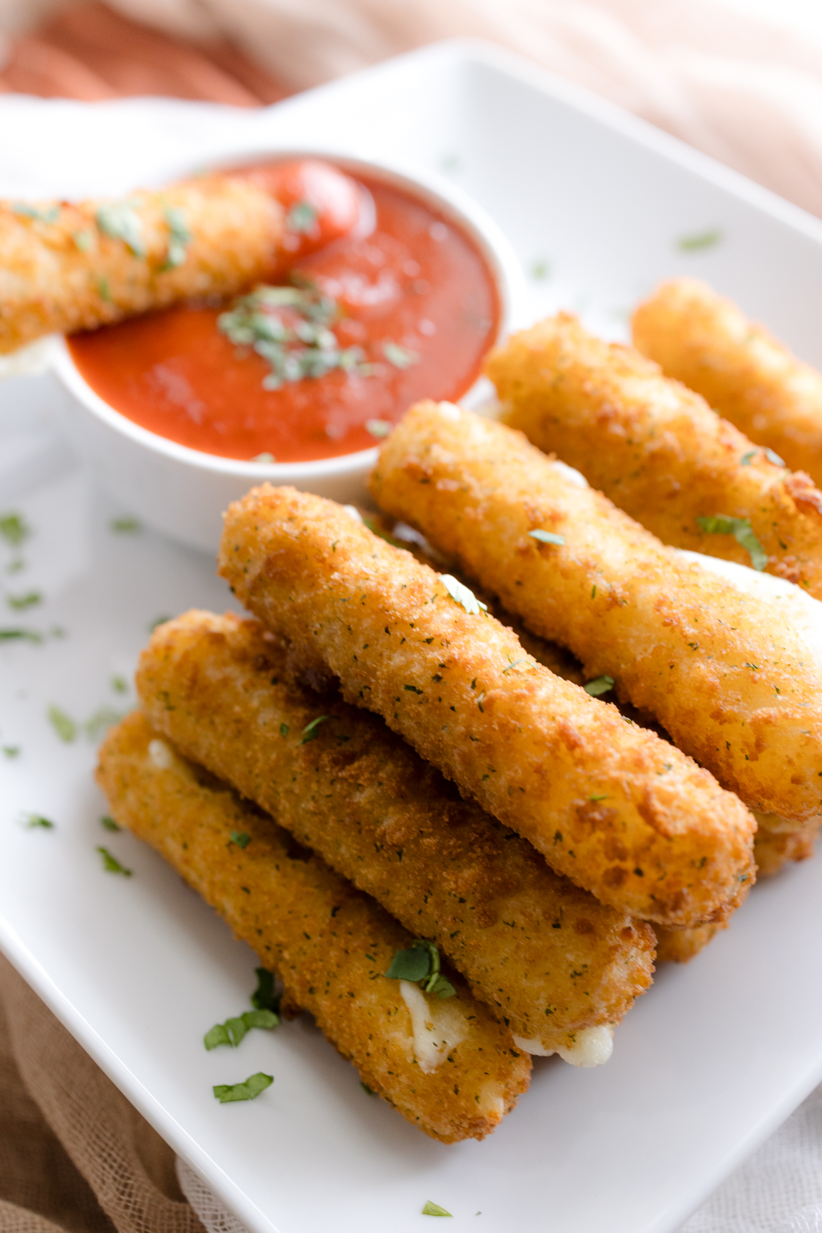 Angled view of Air Fryer Mozzarella Sticks next to a red dipping sauce in a white dipping bowl with a mozzarella stick dipped in it.