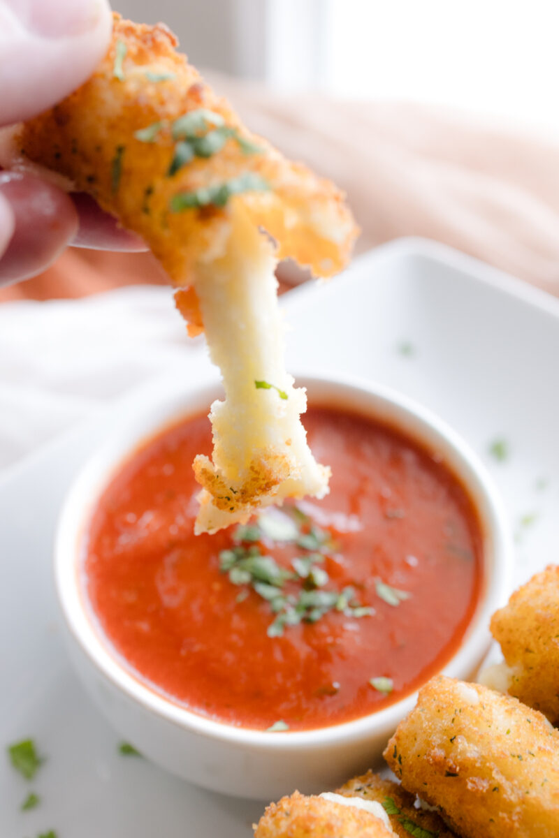 Close up angled view of a hand holding a broken and stringy mozzarella stick in front of a red sauce in a white dipping bowl.