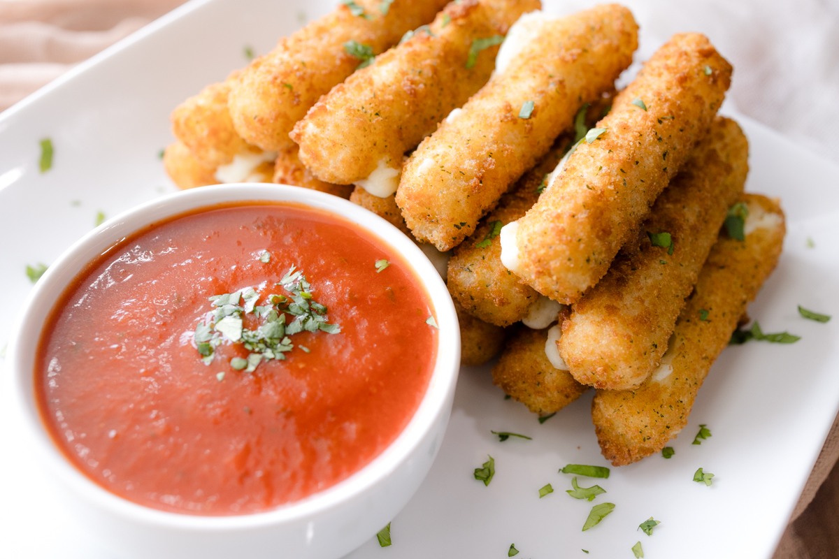 Angled view of Air Fryer Mozzarella Sticks next to a red dipping sauce in a white dipping bowl with a mozzarella stick dipped in it.