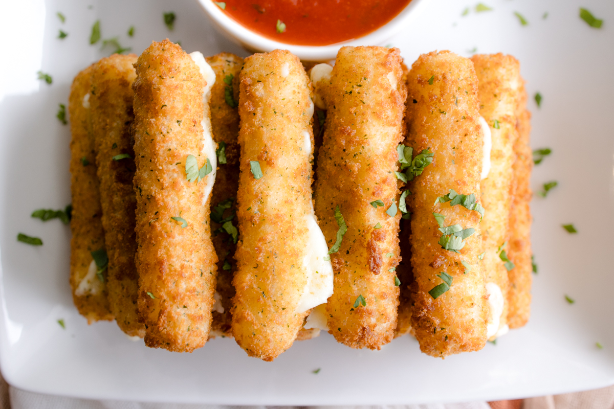 Close up overhead image of Air Fryer Mozzarella Sticks next to a red dipping sauce in a white dipping bowl.