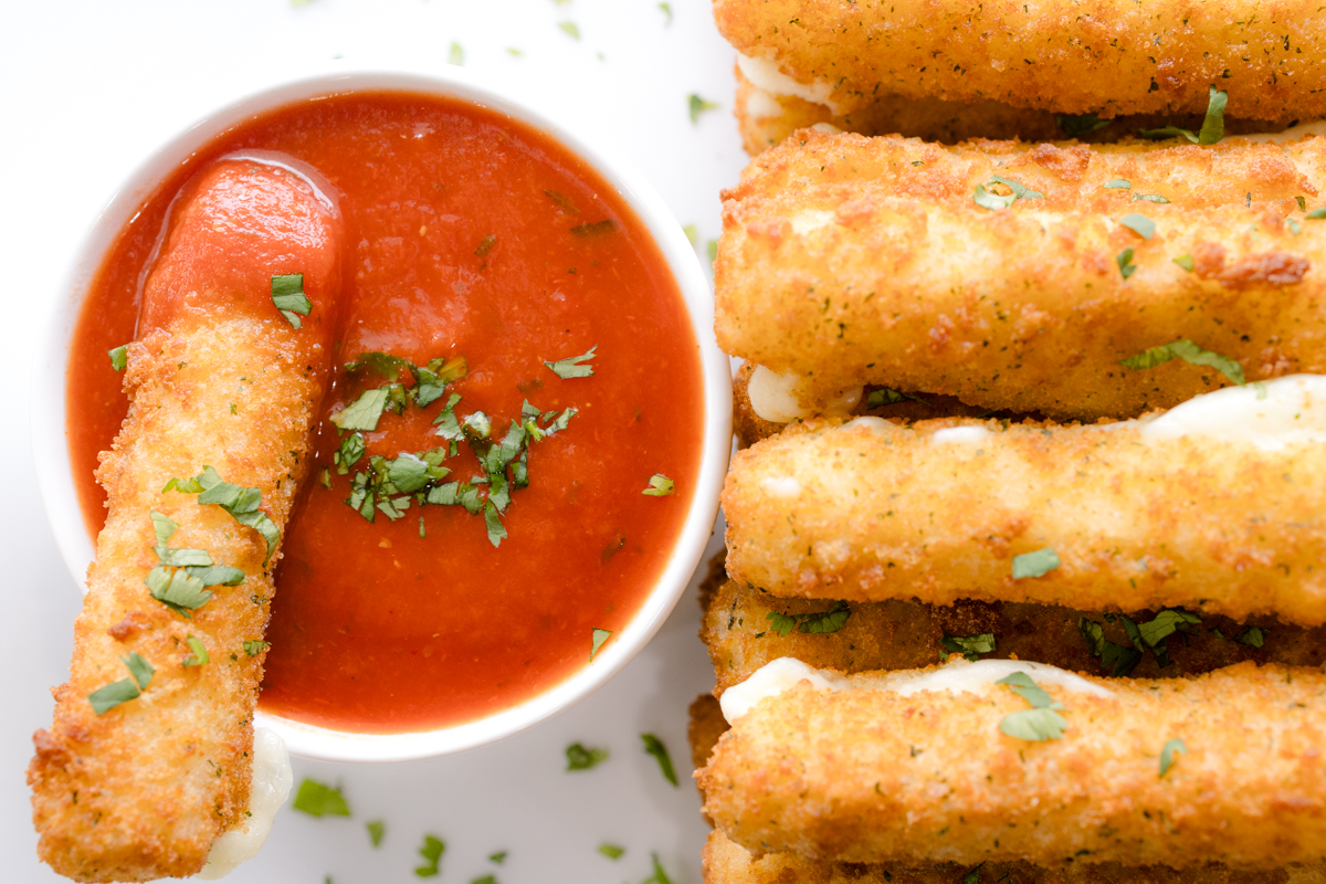Close up overhead image of Air Fryer Mozzarella Sticks next to a red dipping sauce in a white dipping bowl with a mozzarella stick dipped in it.