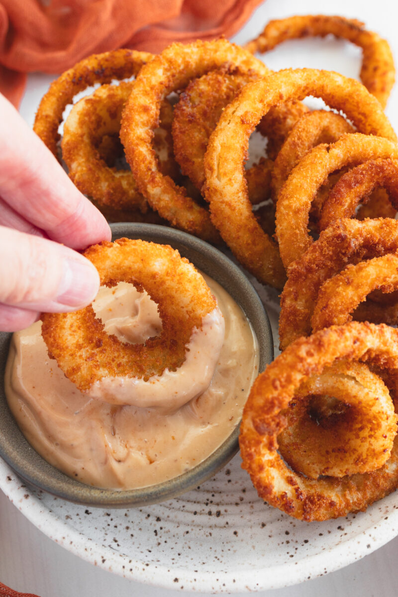 Angled view of a plate of Onion Rings with one being dipped into a dipping sauce.