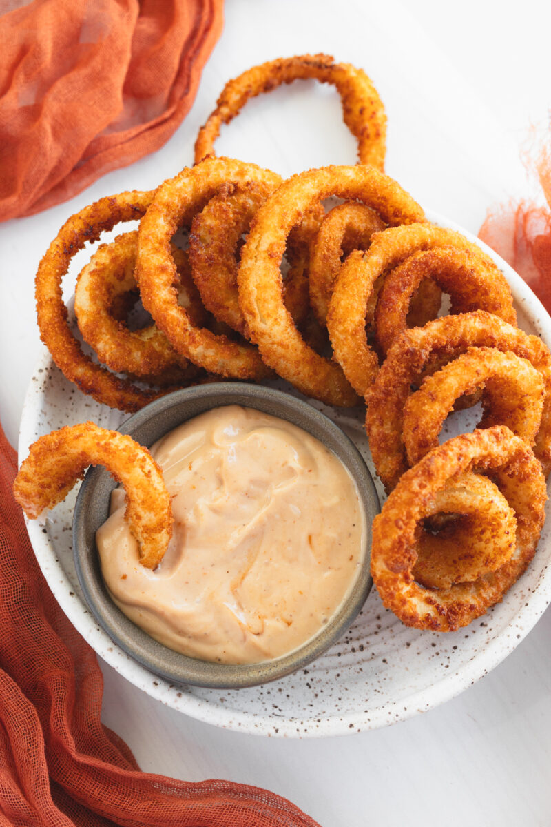 Angled view of a plate of Onion Rings with a dipping sauce.