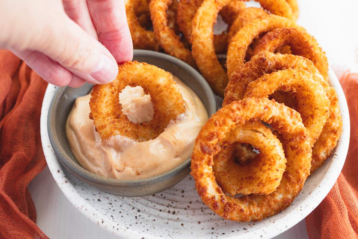 Angled view of a plate of Onion Rings with one being dipped into a dipping sauce.