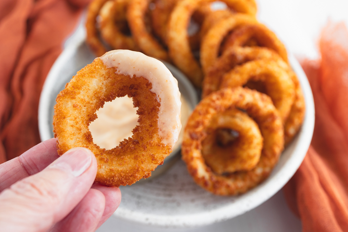 Close up of a hand holding an Onion Ring with sauce on it, over a plate of Onion Rings.