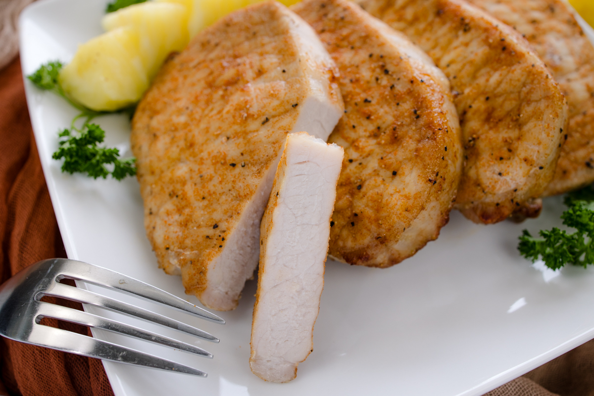 Close-up angled overhead view of a pork chop, sliced to clearly show juicy texture and crispy edges, on a white plate next to a fork and stacked beside other pork chops, with parsley garnish and lemon wedges in the background.