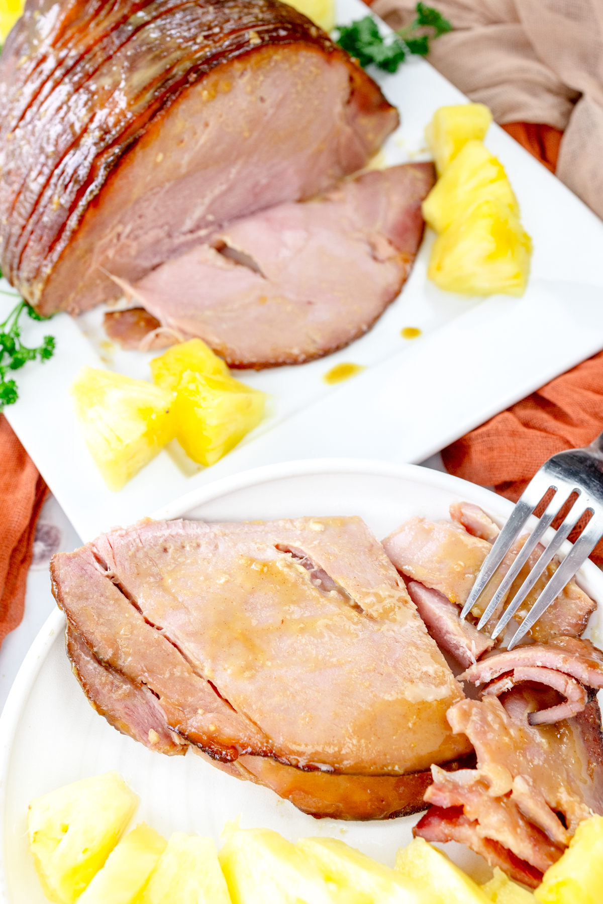 Angled view of Air Fryer Ham slices on a white plate with pineapple chunks. The full ham on another plate in the background.