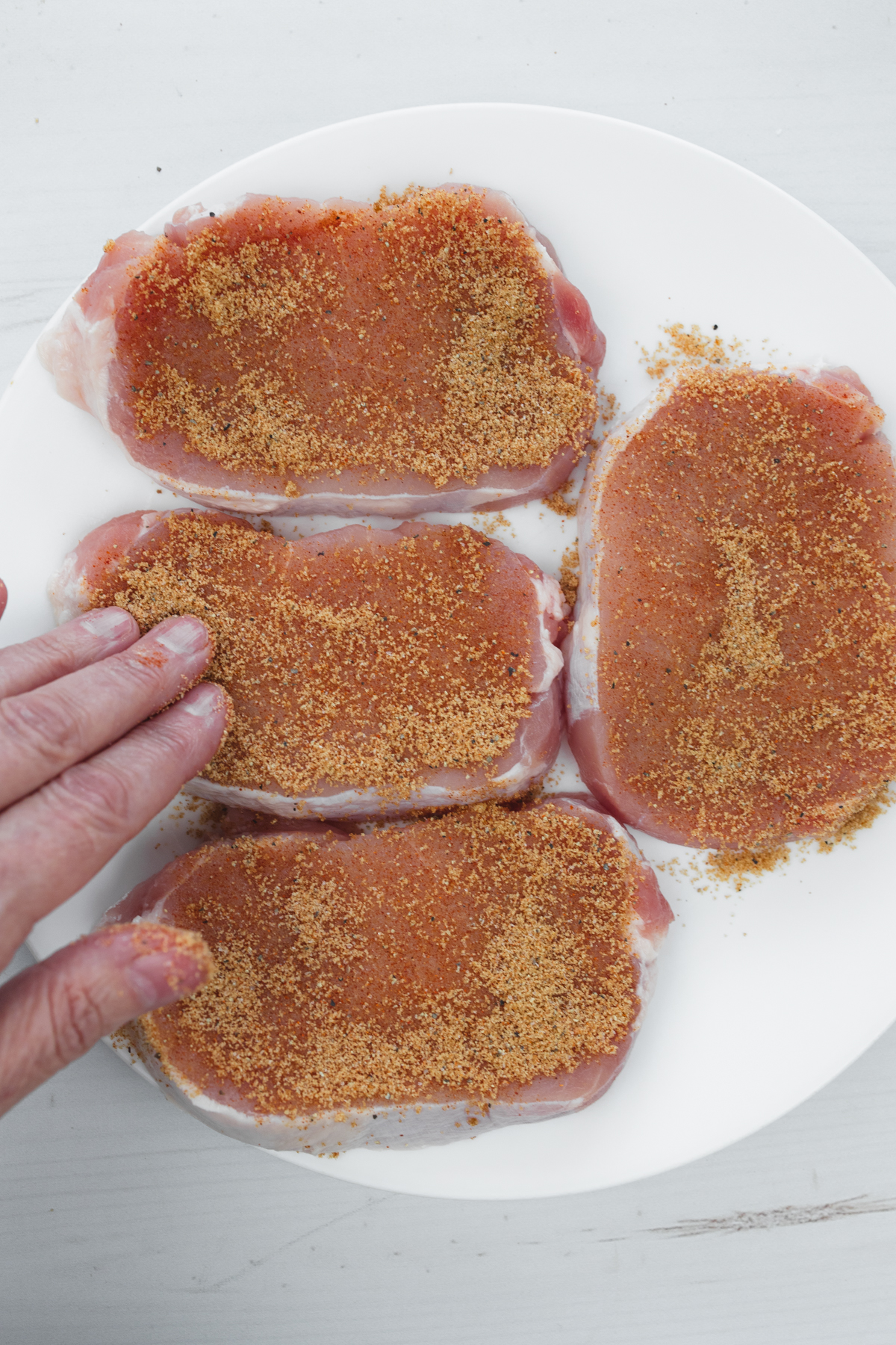 Overhead view of four raw pork chops arranged neatly on a white plate, being seasoned by hand with dry rub.