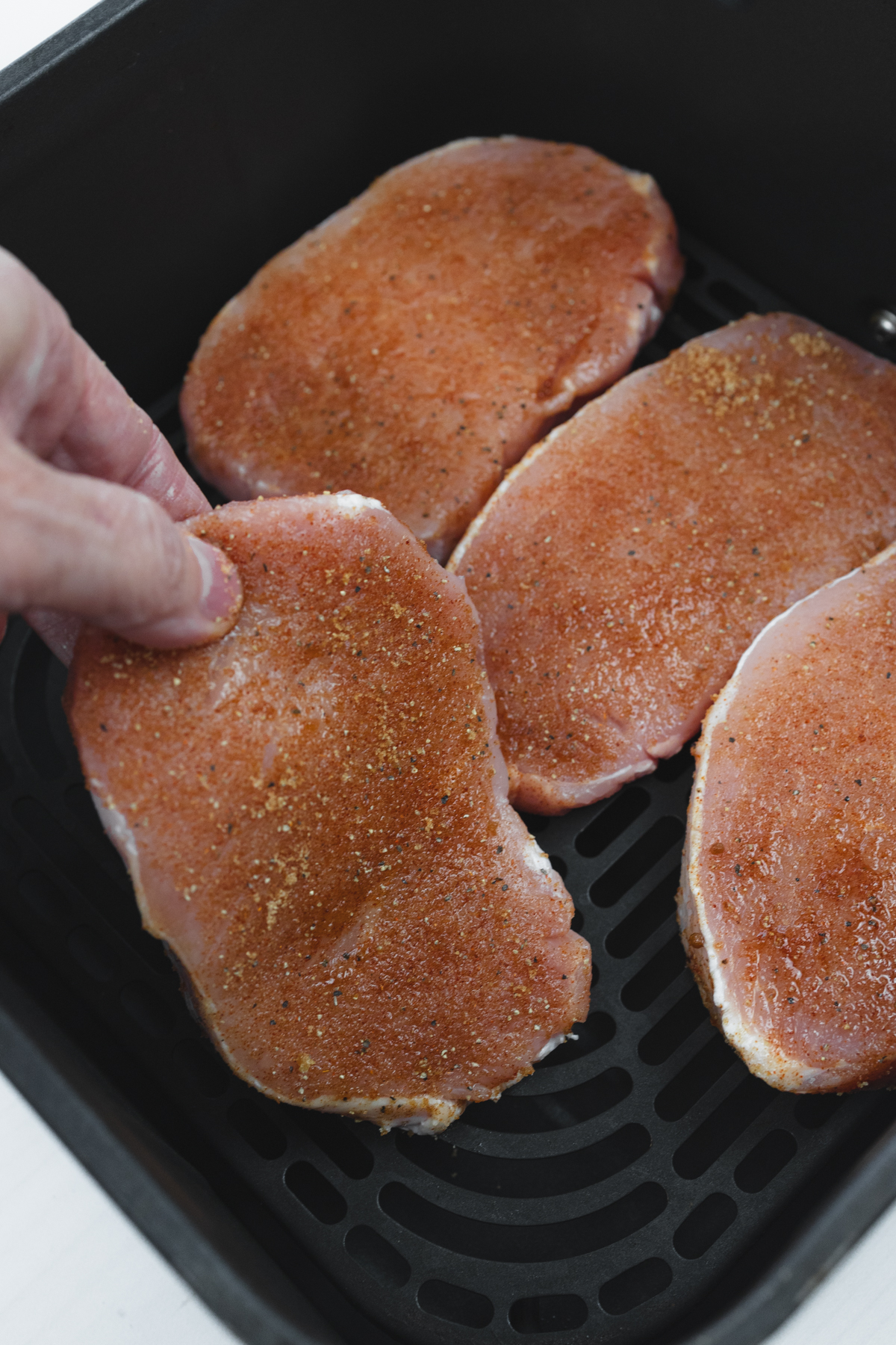 Close-up overhead view of a raw seasoned pork chop being placed into the basket of an air fryer by hand, alongside three other pork chops.