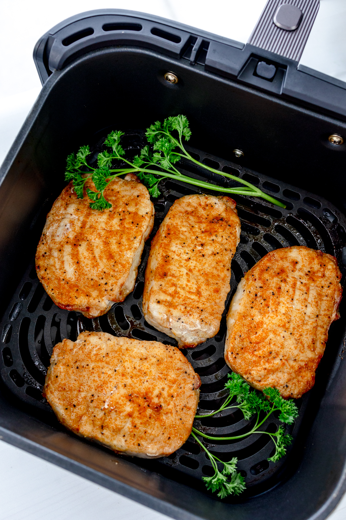 Overhead view of four fully cooked, golden-brown seasoned pork chops in an air fryer basket garnished with fresh parsley.